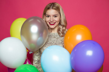 Fototapeta na wymiar Birthday party. Concept of gifts and congratulations. Stylish young smiling beautiful blonde woman in evening dress on a pink background with large multi-colored helium balls.