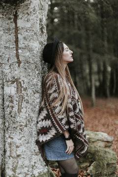 Young woman wearing hat and poncho leaning against tree trunk in autumnal forest