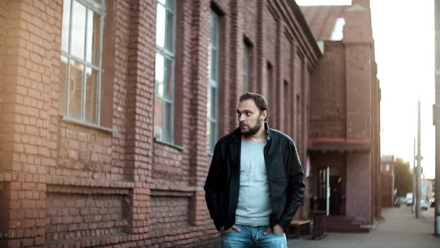 Medium shot portrait of casual European man. Relaxed young male in leather jacket stands waiting. Red brick building 4K.