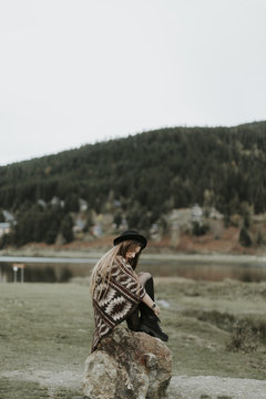 Fashionable young woman wearing hat and poncho sitting on a rock