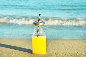 Glass Bottle with Freshly Pressed Tropical Fruits Juice with Straw Standing on Beach Sand. Blue Turquoise Foamy Sea Waves in Background. Golden Sunlight. Vacation Relaxation Freshness Summer