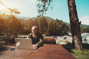 Fototapeta premium Aged grey haired hunter is sitting at big wooden table and checking his e-mail via laptop; mature man is working with his netbook outdoors in nature during his vacation; forester is using his computer