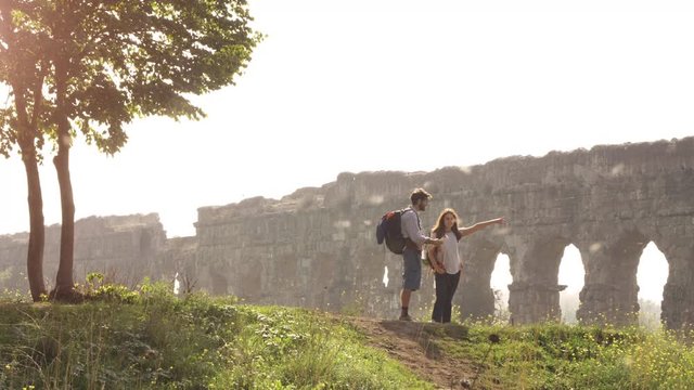 Young lovely couple backpackers tourists reading map pointing directions roman aqueduct arches in parco degli acquedotti park ruins in rome on romantic misty sunrise with guitar and sleeping bag slow