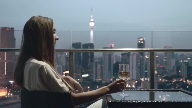Happy woman drinking a glass of white wine while sitting in rooft bar in the city or on the terrace with cityscape at sunset. Vacation,happiness,freedom concept.