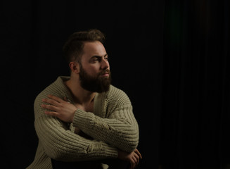 The young model posing in the studio is dressed in a cardigan and is in his hand pockets and bearded ... sitting on a chair, looking ahead.