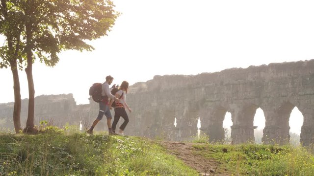 Young lovely couple backpackers tourists walking holding hands arrive on hill in front of roman aqueduct arches in parco degli acquedotti park ruins in rome on romantic misty sunrise with guitar and