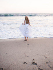 Young girl wearing white dress on beach holding up her dress with toes almost in the ocean