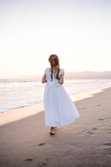 Fototapeta na wymiar Young girl in white dress walking away from the camera in the sand along the waterline of the beach