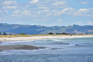 Oceanic surf on flat sandy coast at Maketu estuary with hills in background.