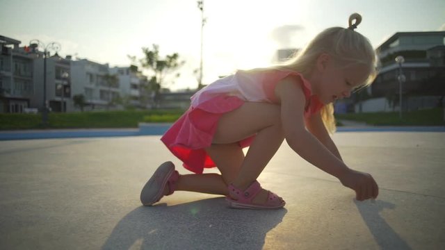 Funny blonde baby girl draws chalk on the playground at sunset rapid slow motion