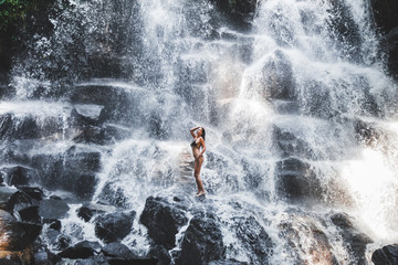 Woman enjoying under stream of big and beautiful cascade waterfall. Girl with slim body and long hair. Kanto Lampo in Ubud area