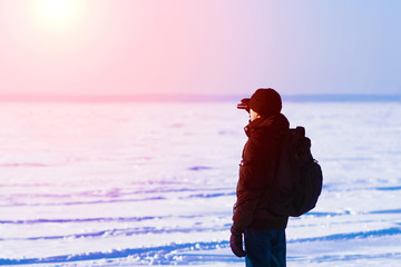 Man in black jacket, knitted hat and blue jeans looking through binoculars at sunset. Rucksack on...