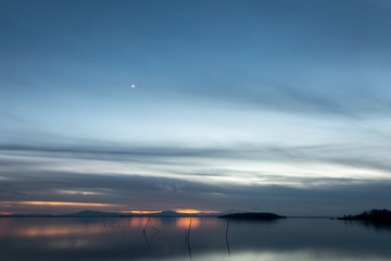 Fototapeta na wymiar Beautiful view of Trasimeno lake (Umbria, Italy) at dusk, with blue and orange tones and moon in the sky