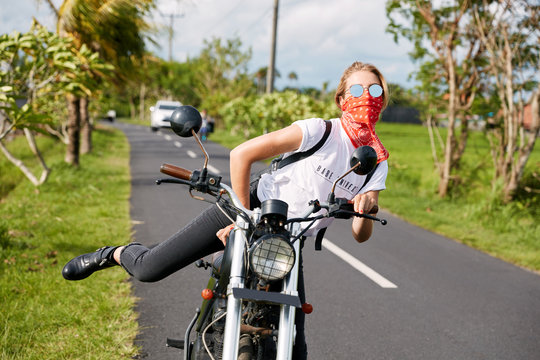 Extremal female biker makes tricks on motorbike, rides on road, coveres mouth with bandana, wears sunglasses, going to make stop. Outdoor shot of active driver being on transport. Freedom concept