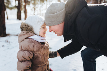 A young happy father with a lovely little daughter is looking at each other cheerfully against the background of a pine winter forest.
