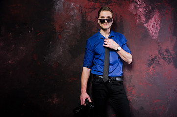 Studio portrait of stylish professional photographer man with camera, wear on blue shirt and necktie, sunglasses.