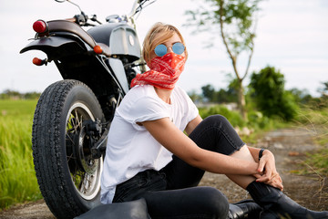 Good looking female dressed casually, wears sunglasses, sits on road near motorbike, rides in countryside at open road, during beautiful sunshine. Time to relax for biker. Active lifestyle concept