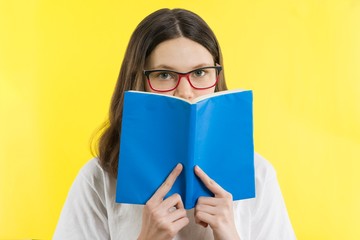 Closeup portrait teenage girl with eye glasses looking over a book, yellow background