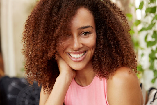 Close up shot of positive dark skinned teenage girl has Afro hairstyle, dressed casually, has shining smile, rests indoor with close friend or boyfriend, being in good mood. People, beauty, ethinicity