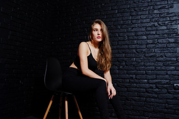 Fototapeta na wymiar Handsome brunette girl wear on black, sitting and posing on chair at studio against dark brick wall. Studio model portrait.