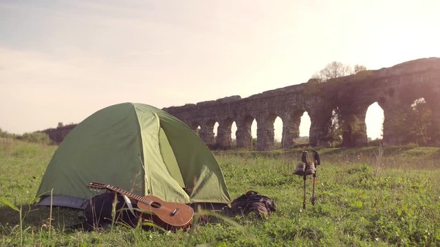 Young man backpacker wakes up exits camping tent in front of roman aqueduct arches in parco degli acquedotti park ruins in rome at sunset sticks in ground slow motion