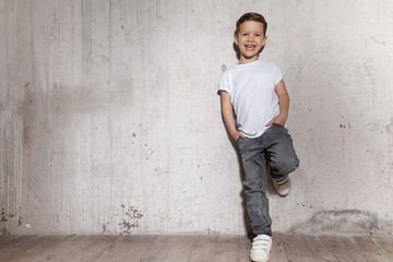 Little fashionable boy posing in front of gray concrete wall. Portrait of smiling child in white T-shirt and gray trousers. Concept of style and fashion for children. © deniskomarov