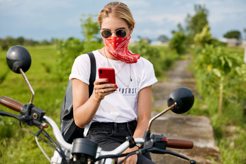 Fototapeta premium Outdoor shot of fashionable young female biker uses smart phone while sits on motorbike, stops to have break after long riding. Hipster girl texts on cell phone, plans her route, uses online map