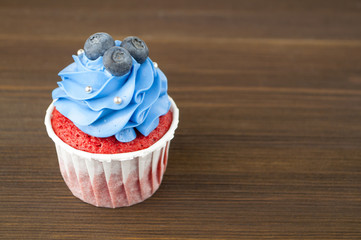 Cupcake red velvet with blue whipped cream decorated with blueberry, silver confectionery balls on dark wood table. Picture for a menu or a confectionery catalog.