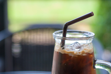 Close up iced espresso coffee with brown straw in crystal high-shape glass on the table at coffee shop with blurred noticable background that it is plant and chair, refreshing drink
