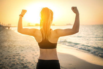 Back view of strong sporty girl showing muscles at the beach during sunset. - obrazy, fototapety, plakaty