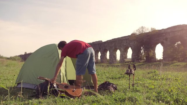 Young man backpacker outside camping tent picks up guitar and walks on grass in front of roman aqueduct arches in parco degli acquedotti park ruins in rome at sunset sticks in ground slow motion