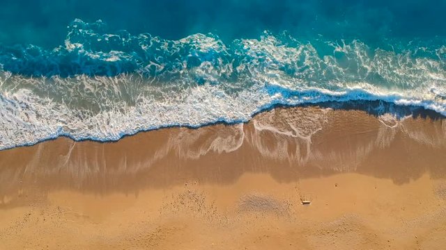 Aerial view looking straight down onto crashing waves hitting a golden beach while panning to the right