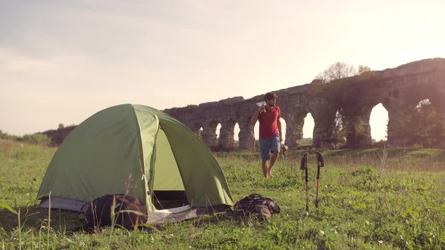 Young man backpacker outside camping tent picks up guitar and walks on grass in front of roman aqueduct arches in parco degli acquedotti park ruins in rome at sunset sticks in ground slow motion