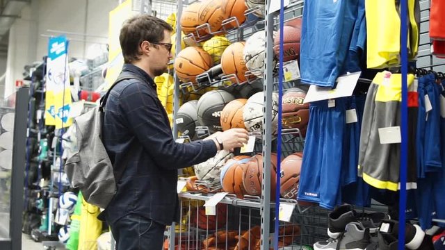 Side view shot of a adult man wearing casual clothes and backpack in sport store. He is choosing new basketball.