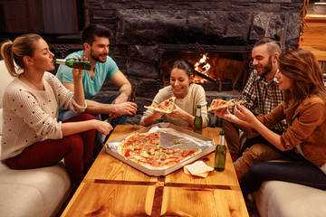 Group of friends having fun at home while toasting beer and eating pizza