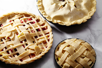 Pie crust design ideas - various ways of pie decoration with lattice and leaves. Apple, strawberry and raspberry pies uncooked on white marble table.