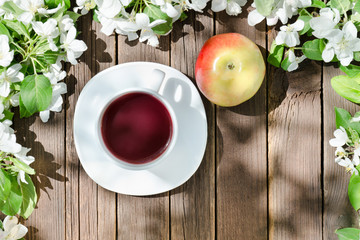 Mug of tea and ripe apple among blossom apple on a wooden background.