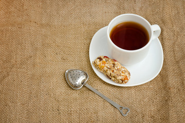 Cup of tea, bar of muesli and tea-strainer on sackcloth. Top view