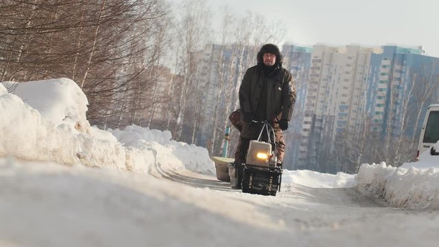 Man in winter clothes riding on mini snowmobile with a trailer and a passenger on a winter road