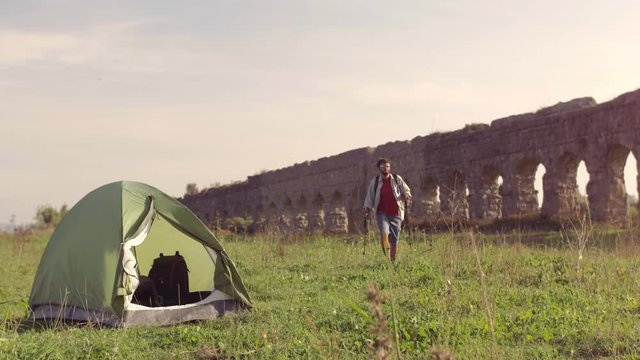 Young man backpacker outside camping tent walks barefeet with sticks on grass in front of roman aqueduct arches in parco degli acquedotti park ruins in rome at sunset slow motion