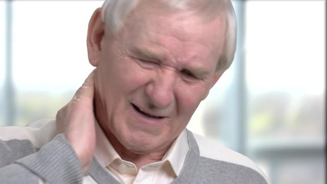 Senior man is having pain in neck. Close up cheerless old man having pain in neck, blurred background.
