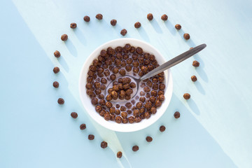 Chocolate cereal balls in white bowl on blue background. Heart shape from flakes. Dry breakfast cereal top view. Concept for a healthy diet.