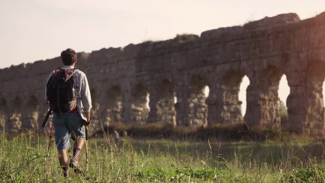 Young man backpacker walks barefeet with sticks on grass in front of roman aqueduct arches in parco degli acquedotti park ruins in rome at sunset slow motion