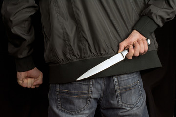 Fototapeta na wymiar hides a big knife behind his back in young man hand of a black jacket and jeans clenched fist. Defend Against a Knife Attack concept close up, selective focus , blurred dark background