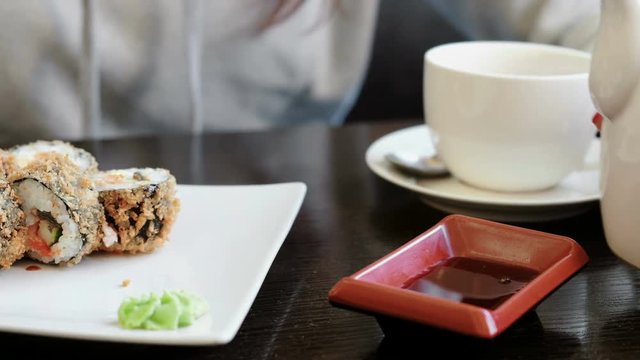 Woman's hand pours soy sauce in small rectangular plate, rolls and cup of tea on background.