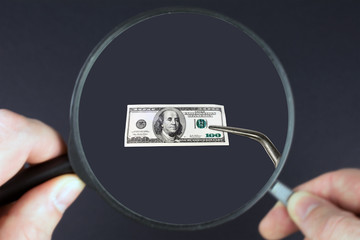 looking through a magnifying glass a small hundred dollar bill held within tweezers, pension plan concept, close up, selective focus, blurred dark background