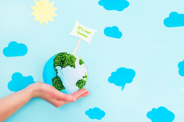 Female Hands holding Earth paper and green sprouts collage model with It's our home pointer on blue background with paper sun and clouds. Earth in your hands, Saving planet concept.