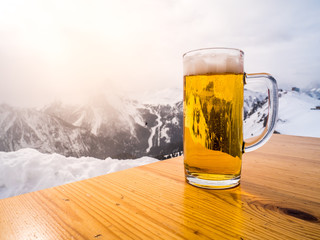 Glass or pint of a beer on a wood table on the top of the alps. Sun light is shining on the winter...
