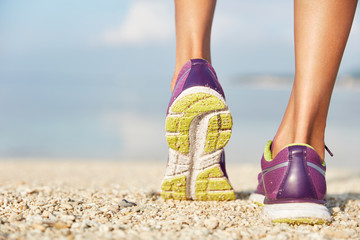 Crropped shot of unrecognizable female in purple sneakers stands on shell beach, wears sport shoes, has morning workout or jogging outdoor, being in movement. Sport and healthy lifestyle concept