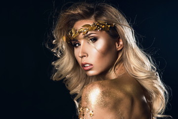 Gold Woman skin close up portrait. Beauty fashion model girl with Golden make up, hair and jewellery on black background. Diadem and bracelet. Metallic, glance art, long blonde shining hair wavy style